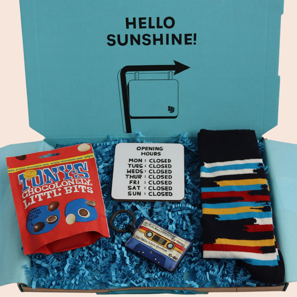 turquoise gift box with the words hello sunshine on containing Tonys chocolate, a coaster with opening hours mon to sun closed, cassette shaped leather keyring, and multi colour stripe socks. The packaging is plastic free with turquoise tissue with turquoise paper shred