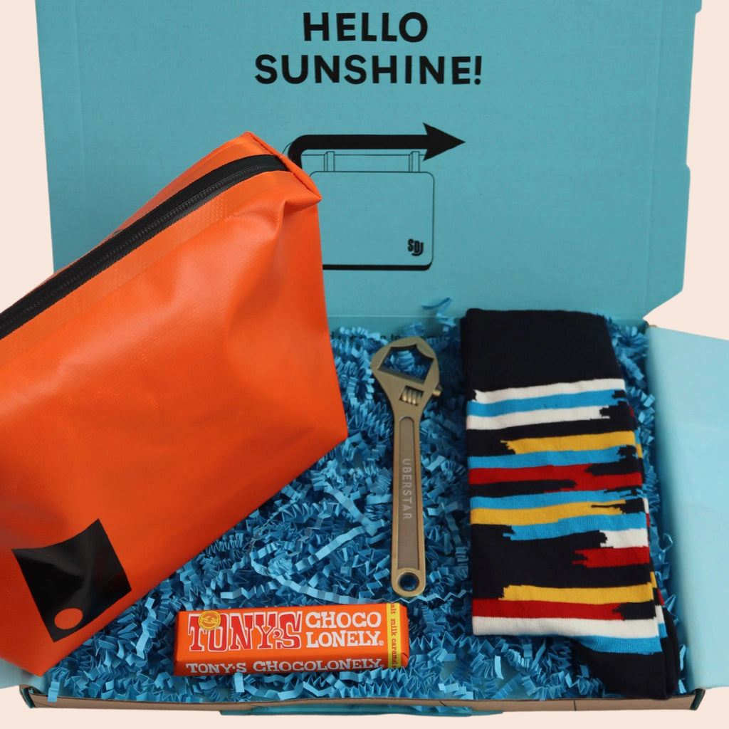 turquoise gift box with the words hello sunshine. The box contains Tonys chocolate, orange dry bag washbag, wrench shaped bottle opener, and multicoloured stripe socks. The packaging is plastic free with turquoise tissue with turquoise paper shred