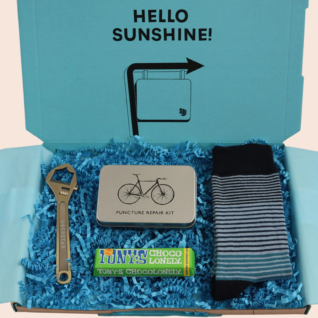 turquoise gift box with the words hello sunshine. The box contains Tonys chocolate, bike puncture repair kit, wrench shaped bottle opener, and blue stripe socks. The packaging is plastic free with turquoise tissue with turquoise paper shred