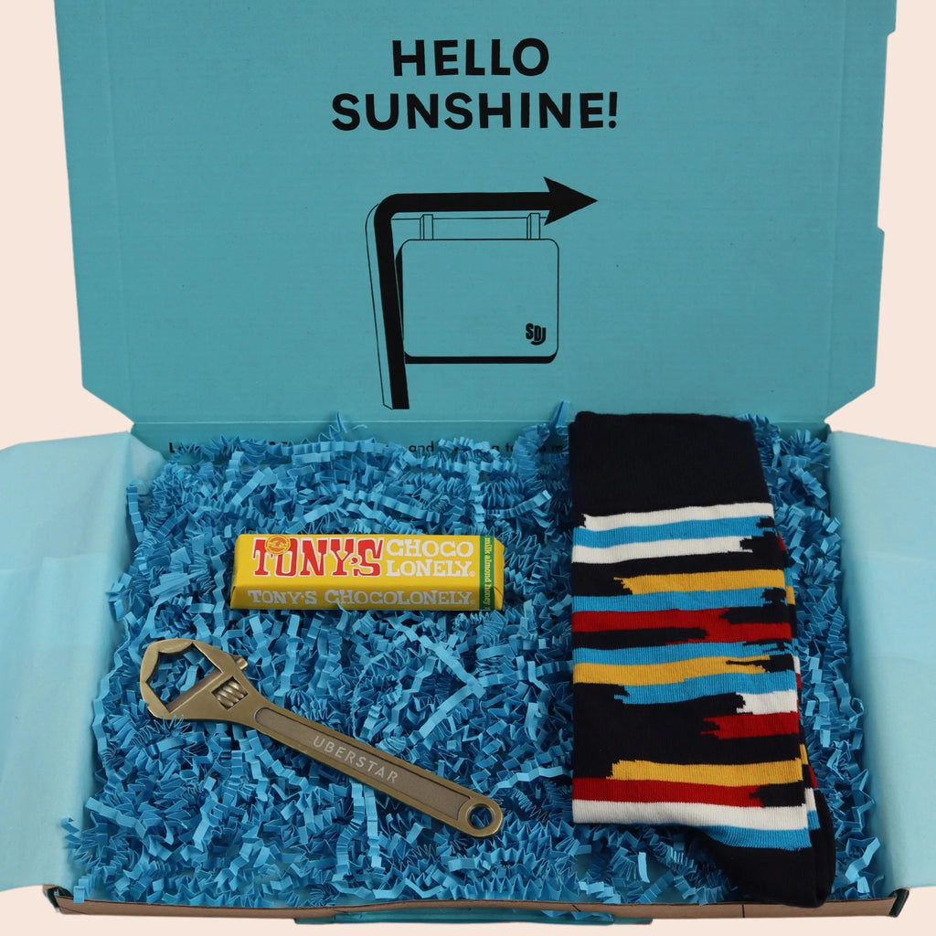 turquoise gift box with the words hello sunshine. The box contains Tonys chocolate, wrench shaped bottle opener, and multicoloured stripped socks. The packaging is plastic free with turquoise tissue with turquoise paper shred